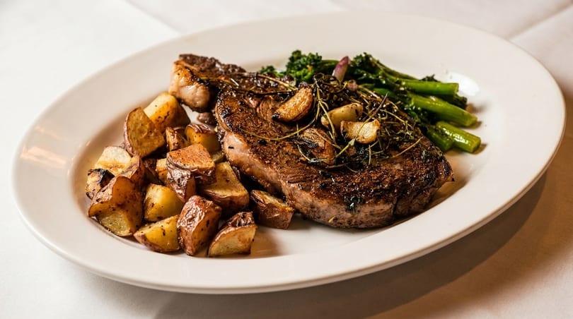 Grilled steak with roasted potatoes, broccolini, garlic, and herbs on a white plate.