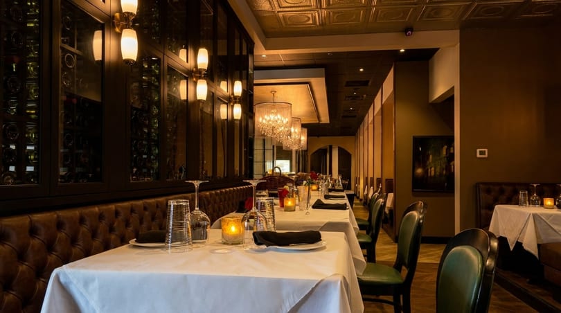 Elegant restaurant interior with white-clothed tables, leather banquettes, wine displays, and chandeliers.