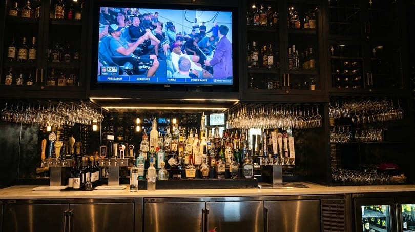 A well-stocked bar with a large TV showing a sports game, liquor bottles, and hanging glassware.