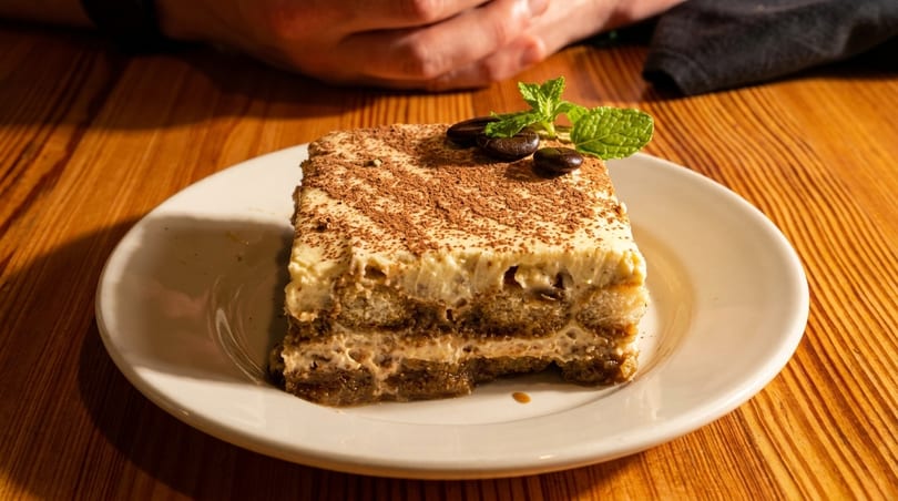 A slice of tiramisu with cocoa powder, coffee beans, and mint on a white plate.