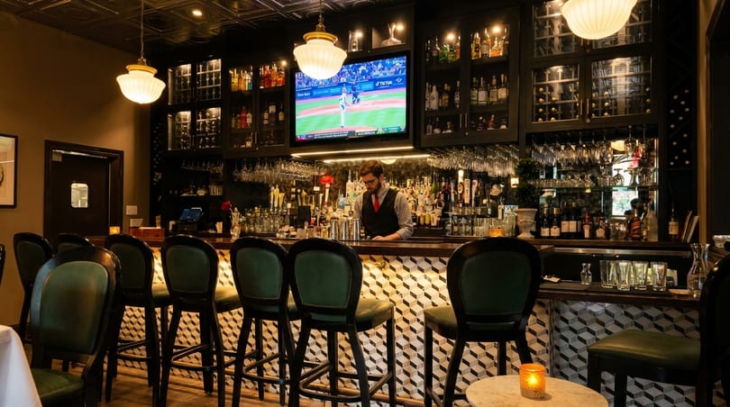 A bartender works behind a well-stocked bar with green stools and a TV showing baseball.