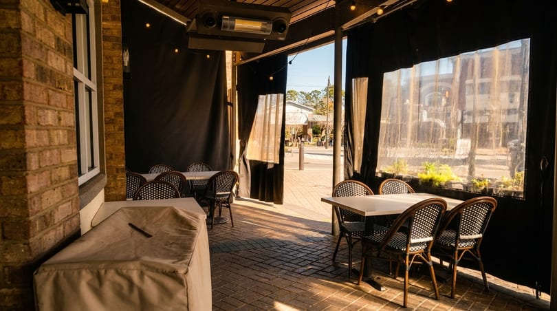 Outdoor patio with brick walls, black curtains, bistro tables, and chairs, with sunlight streaming in.
