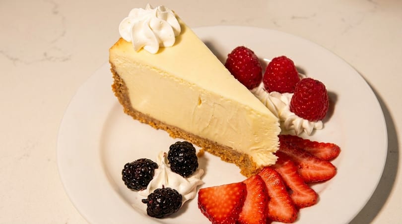 Slice of cheesecake with whipped cream, raspberries, blackberries, and sliced strawberries on a white plate.
