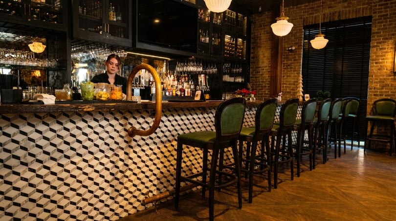 A bartender stands behind a stylish bar with geometric tile, green stools, and brick walls.